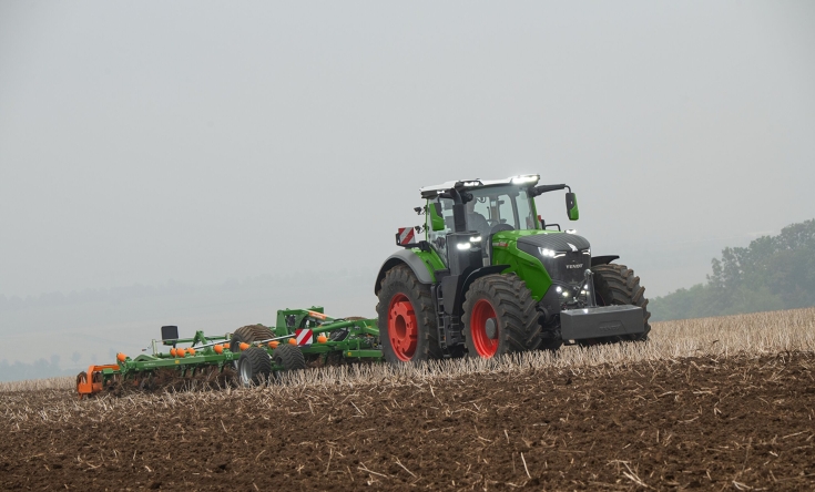 Vihreä Fendt 1000 Vario -traktori muokkaa peltoa vihreällä ja oranssilla Amazone-kultivaattorilla, hämärässä ajovalot ovat päällä.