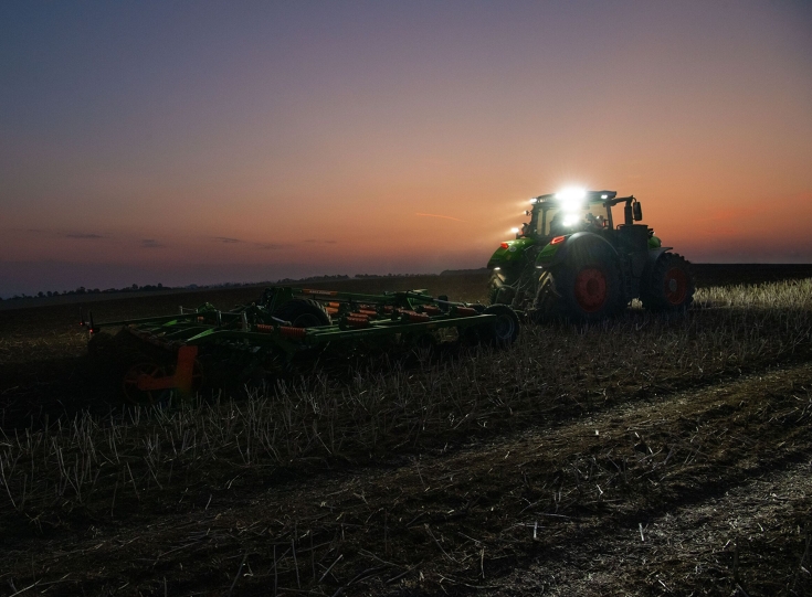 Vihreä Fendt 1000 Vario -traktori kuvattuna takaa muokkaa peltoa vihreällä ja oranssilla Amazone-kultivaattorilla. Kaikki ajovalot ovat päällä, taivaalla näkyy auringonlaskun viimeiset rippeet.