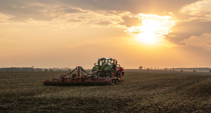 Vihreä Fendt 1000 Vario ajaa Kverneland-etutankin ja lautasäkeen kanssa pellolla kohti auringonlaskua