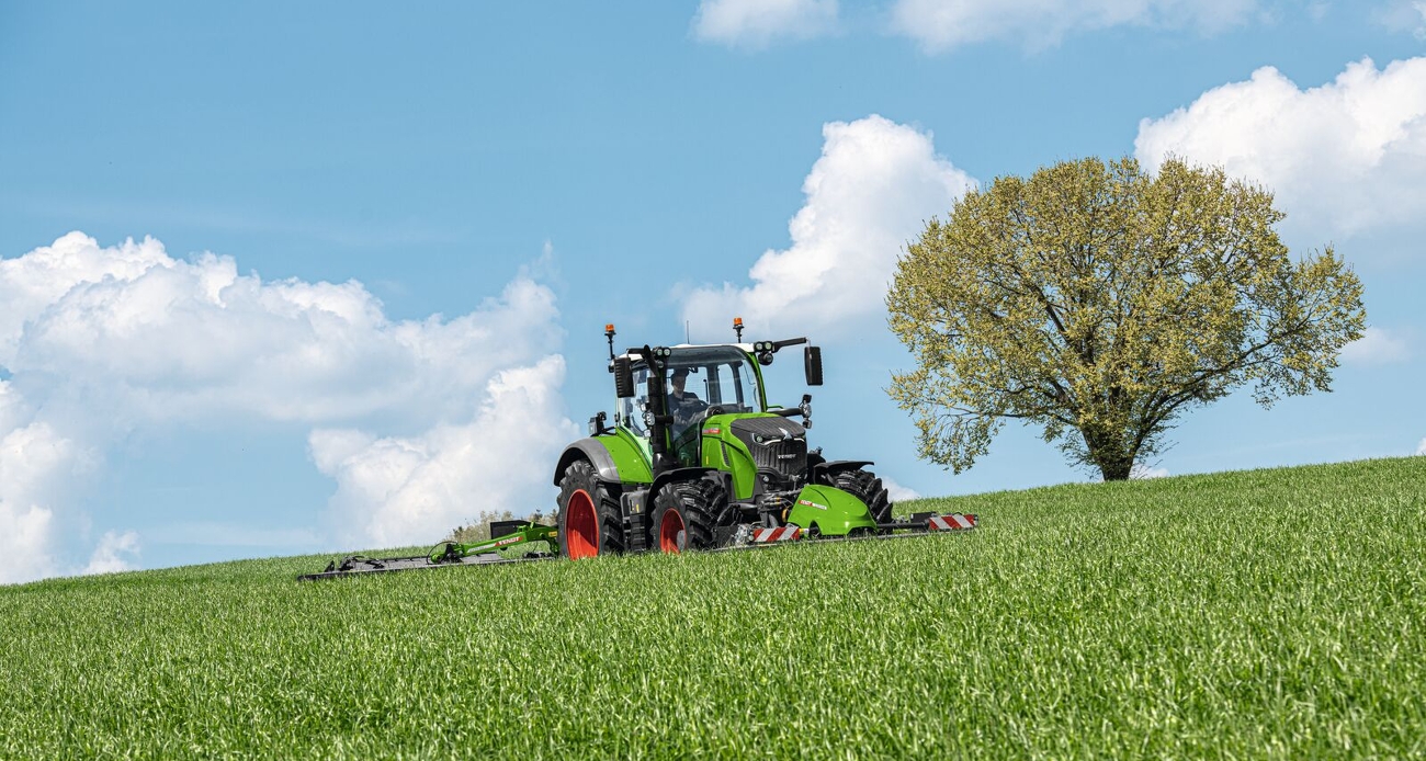 Vihreä Fendt 700 Vario Gen7 niittää Fendtin etu- ja takaniittokoneella niittyä, taustalla näkyy puu.