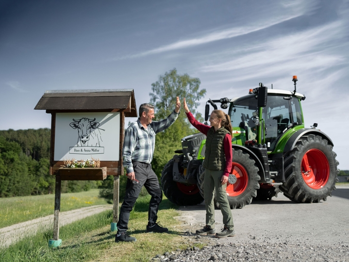 Maanviljelijä ja tytär seisovat maatilakyltin edessä, Fendt 500 Vario taustalla
