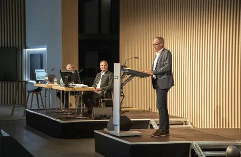 Vue latérale du Dr Wolfgang Hell à un pupitre avec le logo Fendt, en train de prononcer son discours. À l'arrière-plan, on aperçoit deux autres personnes assises à des tables avec des ordinateurs, qui l'écoutent.