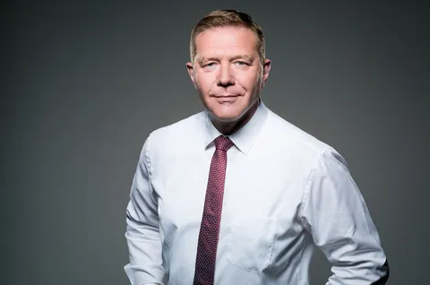 Portrait de Christoph Gröblinghoff  en chemise blanche et cravate rouge devant un fond gris foncé.