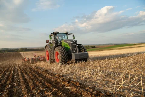 un tracteur Fendt 1000 Vario vert travaille dans un champ de chaumes avec un déchaumeur Horsch rouge, en arrière-plan, on peut voir des champs et une forêt.