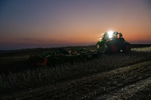 prise de vue de l’arrière d’un tracteur Fendt 1000 Vario vert travaillant dans un champ de chaumes avec un déchaumeur Amazone vert et orange Tous les phares sont allumés, dans le ciel on voit les derniers instants d'un coucher de soleil.