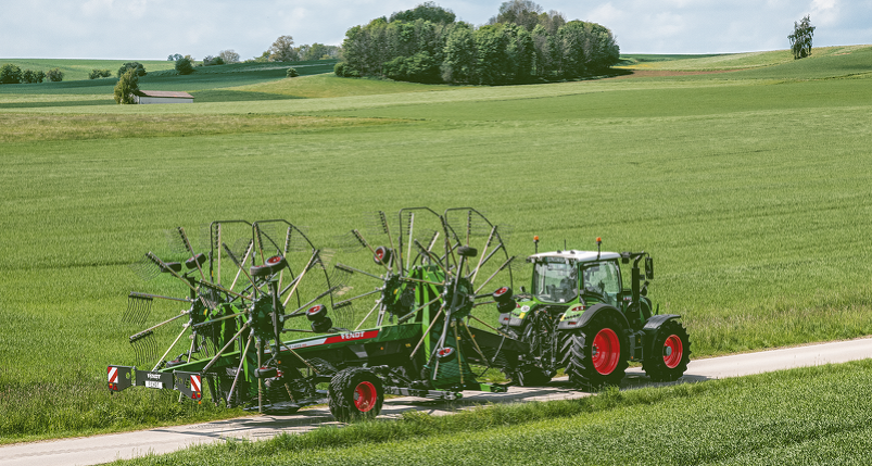 Un tracteur Fendt vert tire une Fendt Former vert-gris avec des andaineurs relevés sur un chemin de campagne pour aller travailler dans les champs.