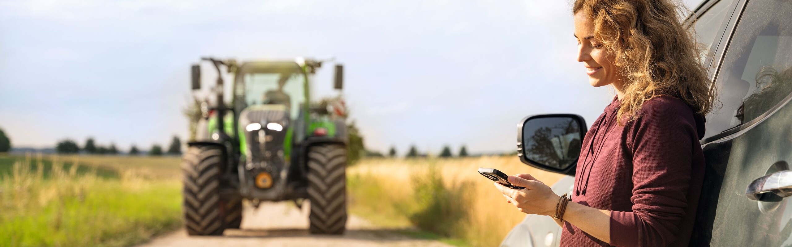 Une femme aux cheveux blonds bouclés et vêtue d'un pull rouge foncé est appuyée contre une voiture et regarde son téléphone portable. Un tracteur Fendt est visible en arrière-plan.