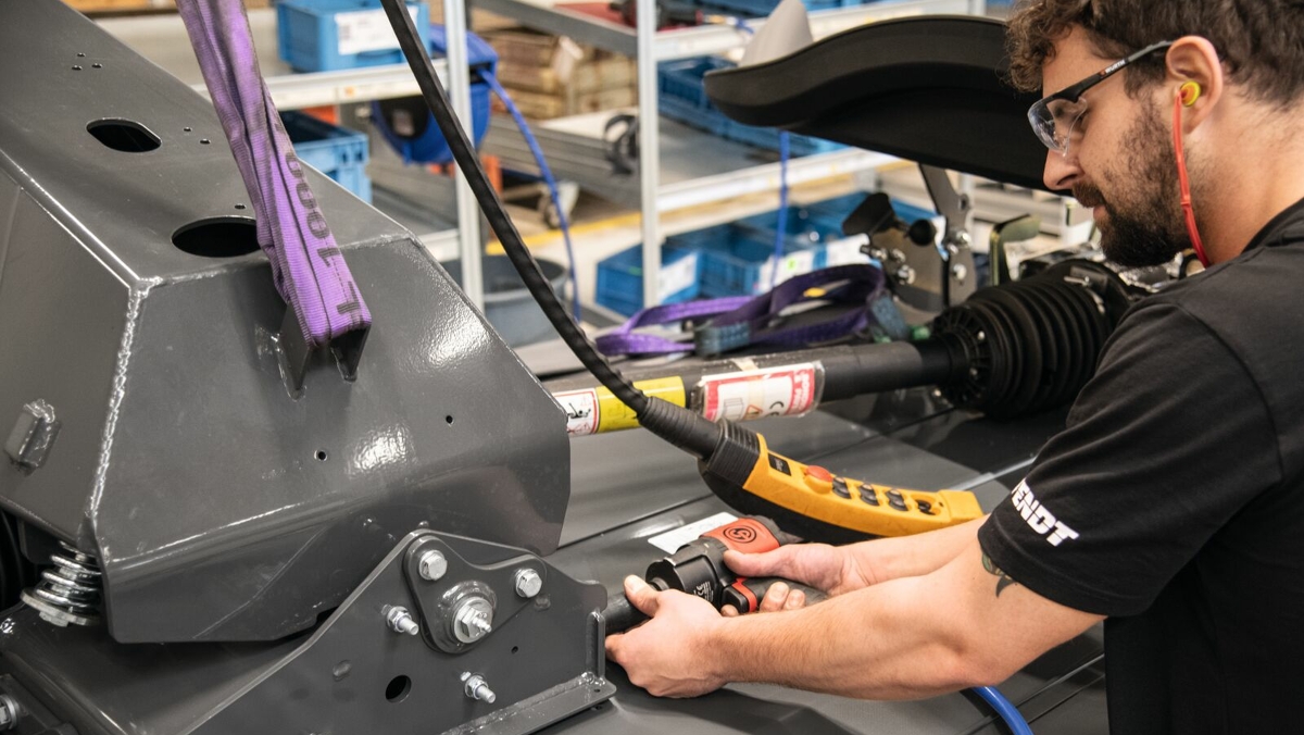 Un employé, vêtu d'une tenue de travail Fendt noire et portant des lunettes de protection, est en train de monter quelque chose sur la chaîne de montage.