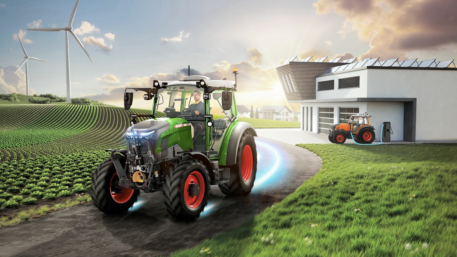 Un Fendt e100 Vario roule sur un chemin rural à proximité d'une localité, tandis qu'à l'arrière-plan, des éoliennes tournent et qu'un autre Fendt e100 se recharge.