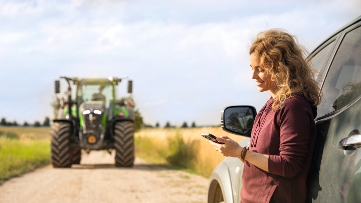 Une agricultrice à la recherche d'un revendeur sur son smartphone. Une agricultrice se tient sur un chemin de campagne, appuyée contre sa voiture, à la recherche d'un revendeur Fendt sur son smartphone. Un tracteur Fendt est visible à l'arrière-plan.