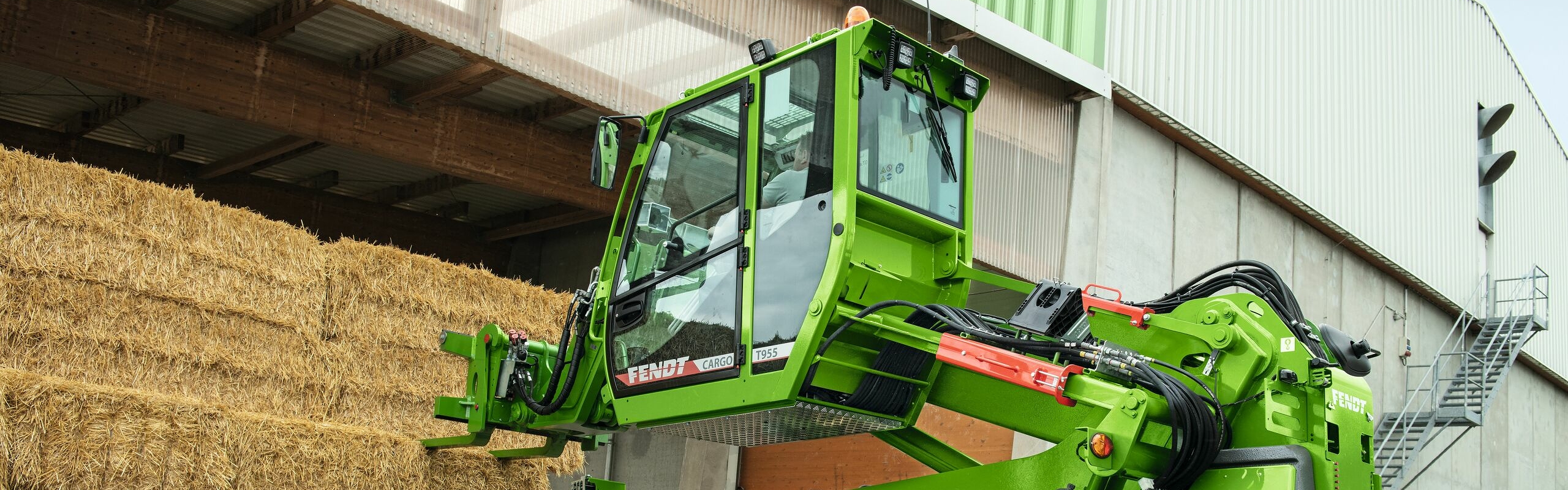 Gros plan latéral sur un agriculteur assis dans une cabine de Fendt Cargo T955 qui s’apprête à saisir une balle de paille avec son godet.