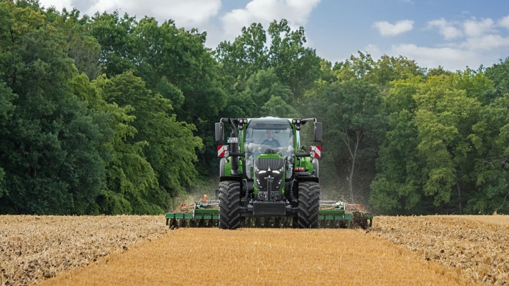 Un Fendt 600 Vario vert en train de travailler le sol dans un champ avec une forêt en arrière-plan