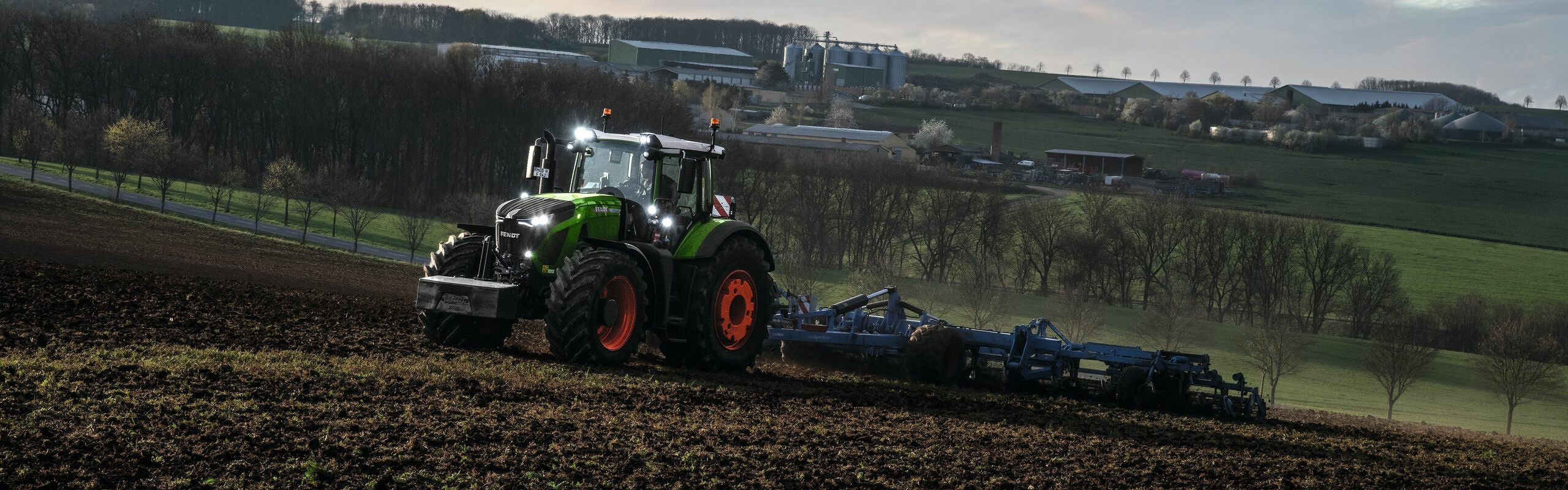 Le tracteur vert Fendt 900 Vario avec des jantes rouges et l’outil porté bleu de Lemken en action dans un champ au crépuscule.