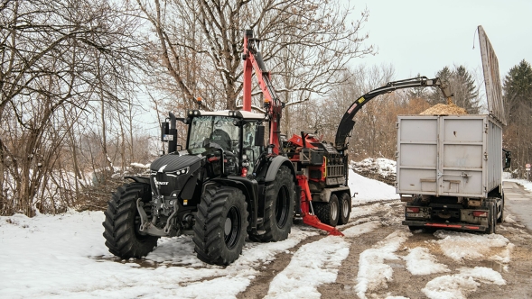 Un Fendt 900 Vario noir avec un outil porté et remorque autochargeuse en train de broyer du bois dans un champ enneigé, entouré d’arbres dénudés en hiver.