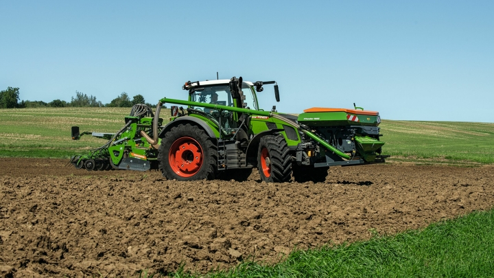 Le Fendt 600 Vario vert avec des jantes rouges et un semoir comme outil de travail se déplace dans un champ. En arrière-plan, on peut voir une prairie verte et un ciel bleu.