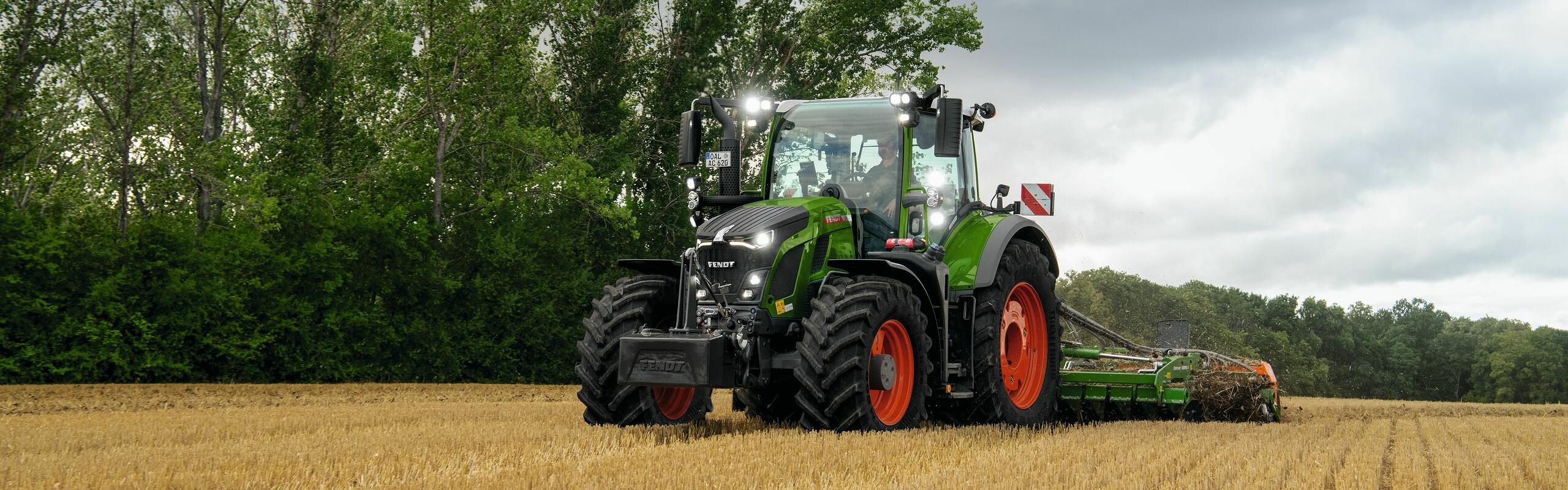 Le tracteur Fendt 600 Vario vert avec des jantes rouges en action dans un champ avec des arbres en arrière-plan et un ciel nuageux.