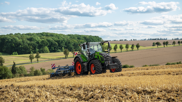 Un tracteur Fendt vert avec des jantes rouges en train de travailler le sol dans un champ récolté avec un paysage de collines en arrière-plan.