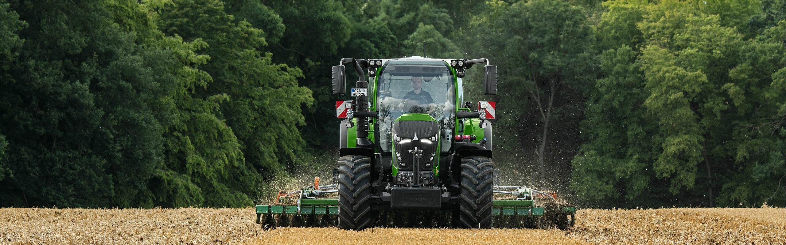 Le Fendt 600 Vario vert est en action avec un outil dans un champ marron. En arrière-plan, une forêt vert foncé et un ciel couvert de nuages.
