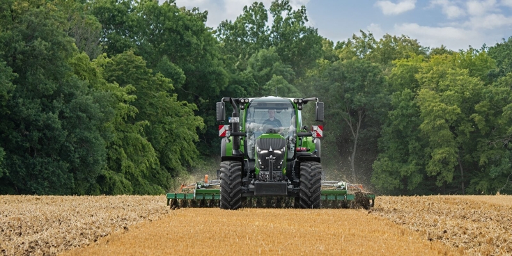 Un Fendt 600 Vario vert en train de travailler le sol dans un champ avec une forêt en arrière-plan