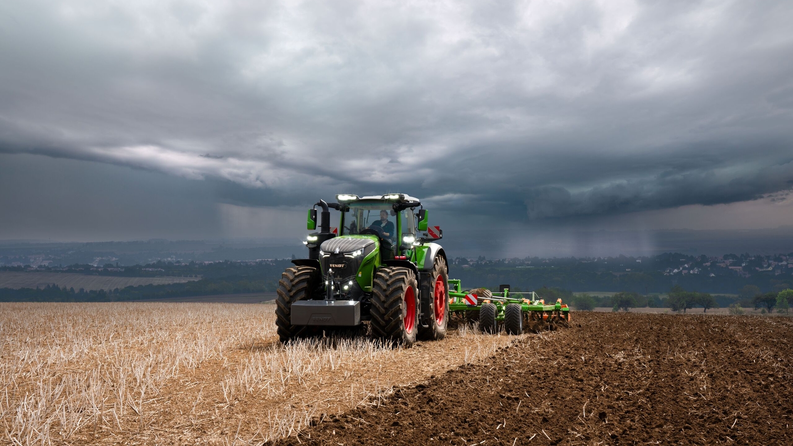 Le tracteur Fendt 1000 Vario effectue encore son travail à pleine puissance dans les champs malgré les lourds nuages d'orage qui s'approchent au loin.