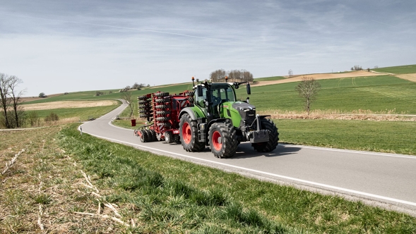 Le Fendt 700 Vario Gen7.1 avec un équipement de transport sur la route dans un environnement rural. Un Fendt 700 Vario Gen7.1 avec un outil attelé roule sur une route de campagne à travers un paysage vallonné au printemps.