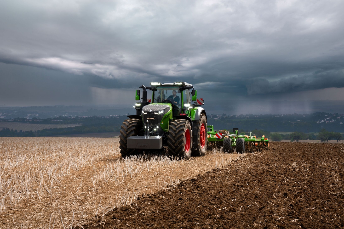 Le tracteur Fendt 1000 Vario effectue encore son travail à pleine puissance dans les champs malgré les lourds nuages d'orage qui s'approchent au loin.