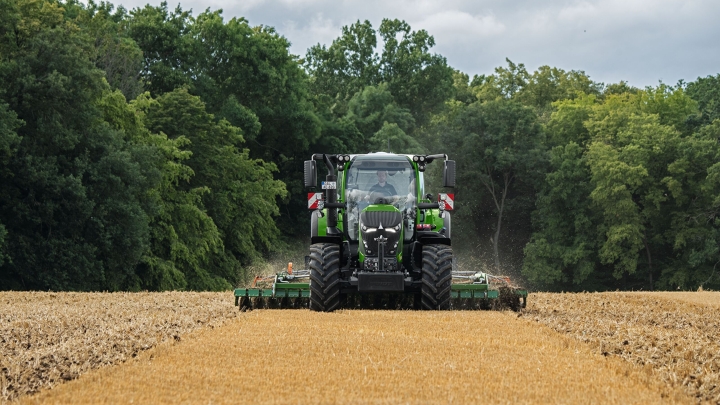Le Fendt 600 Vario vert est en action avec un outil dans un champ marron. En arrière-plan, une forêt vert foncé et un ciel couvert de nuages.