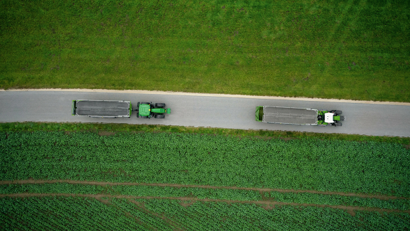 Egy Fendt 700 Vario Gen7 egy rendfelszedő pótkocsival egy ugyancsak rendfelszedő pótkocsit vontató John Deere traktor előtt halad.