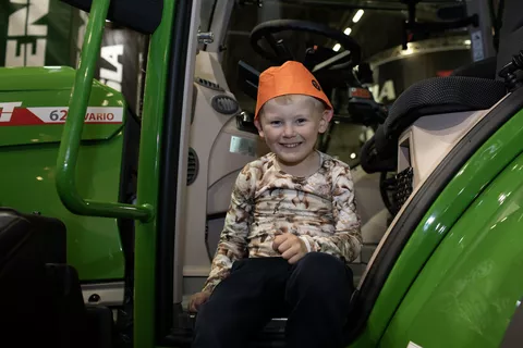 A little boy sits on the steps of a Fendt tractor, smiling happily and wearing an orange hat