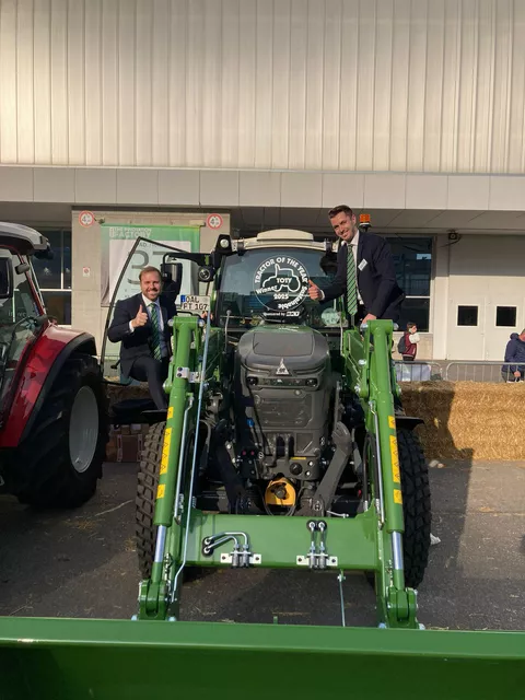 Two Italian Fendt employees sit on the left and right of the Fendt e100 Vario with the TotY 2025 logo on the windscreen and look delighted