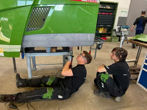 Two men (apprentices) are working on a tractor. One of them is sitting under the bonnet with a hand drill, while the other is crouching down and watching