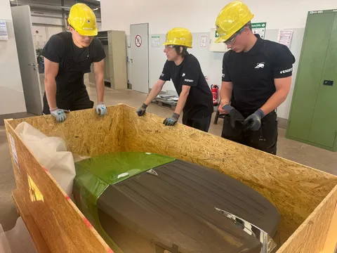 3 men (apprentices) are standing around an open box containing the Fendt bonnet, examining it and talking.