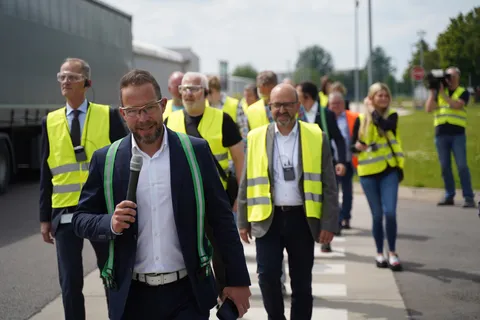 People are walking across a crosswalk on the factory premises and listening to Alexander Schenk speaking into a microphone.