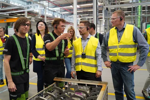People are standing in a production hall wearing yellow reflective safety vests while one of the two apprentices explains something to them.