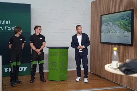 Apprentices and another man are standing in front of a wall. The two apprentices are looking at the man in the suit while he is speaking