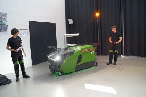 Two men (apprentices) stand next to the green Fendt dispensing station, one on the left and one on the right. The front lights, underbody lighting and rotating beacon are illuminated.