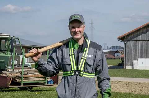 A man in work clothes smiles at the camera while holding a sledgehammer. Behind him is a wooden house (right) and a green Fendt tractor (left) loaded with wooden poles
