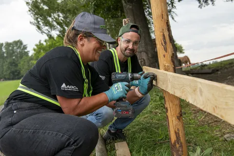 2 people in protective work clothing, one of them a woman (front), squatting in front of a wooden fence. The woman is drilling into the fence while the man next to her watches in the background.