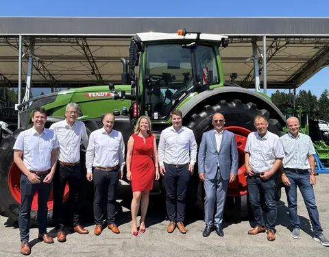 Group photo of 8 people, including 7 men and 1 woman in the middlewith a red dress, in front of a green Fendt tractor.