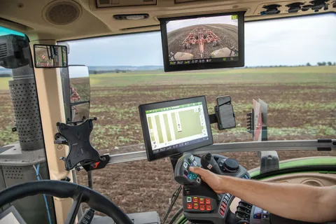 A tractor cab from inside with steering wheel and FendtONE armrest for tractor operation including terminal display for a guidance system. Another screen is anchored in the cab roof, which shows the tractor rear and cultivator via a camera, and a field ca