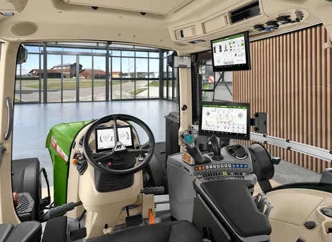 An interior image of the tractor cab from the driver's point of view with steering wheel, FendtONE control armrest with joystick and terminal, and an additional terminal in the roof. Through the front window you can see a hall with a large window and a co