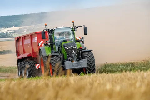 A green tractor pulls a red trailer through a field of golden wheat, kicking up dust.