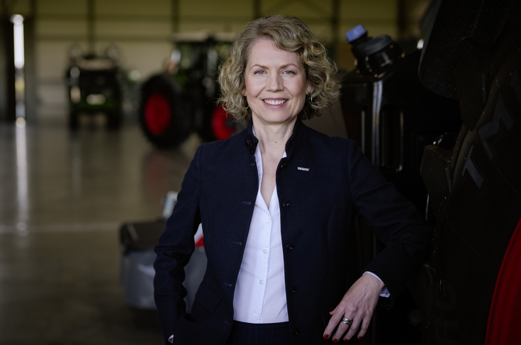A woman with chin-length, curly blonde hair wears a black blazer and a white blouse; Fendt machines can be seen in the background