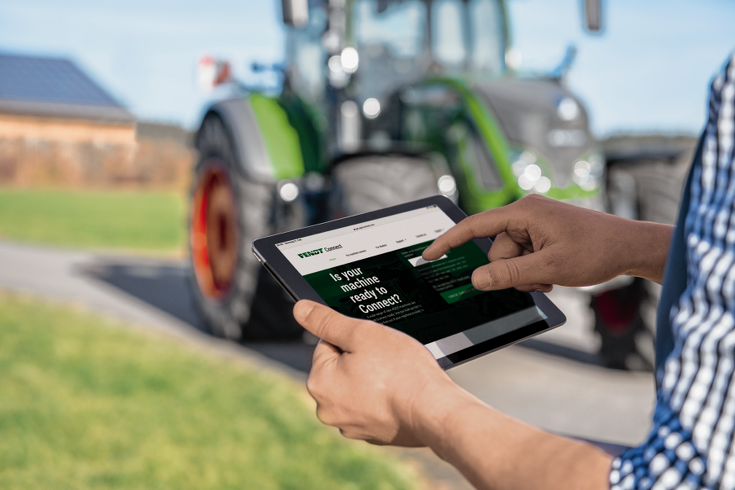 In front of a Fendt tractor one sees a man holding a tablet that shows the review of his machines for Fendt Connect.