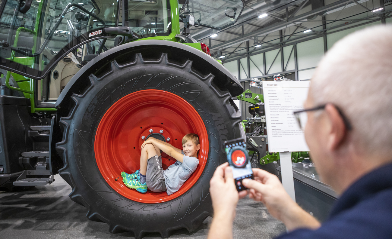 Close-up view of a Fendt tractor, a man is photographing a little boy lying in the tractor’s tyre.