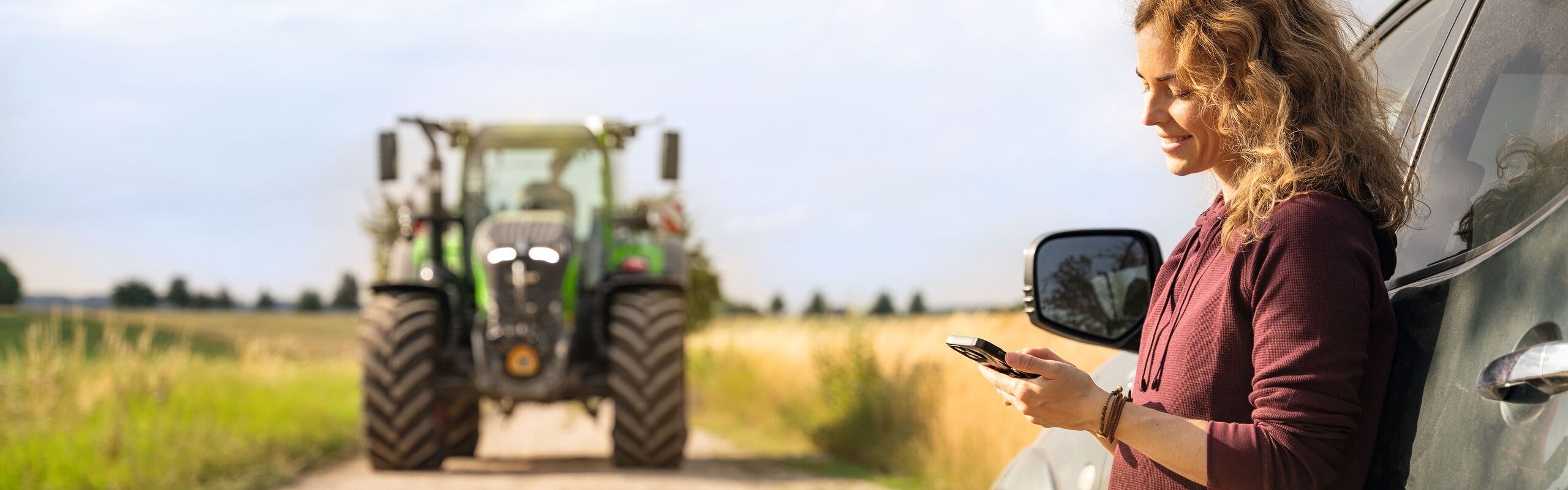 A woman with blonde curls and a dark red jumper standing and leaning against a car and looking at her mobile phone; a Fendt tractor can be seen in the background