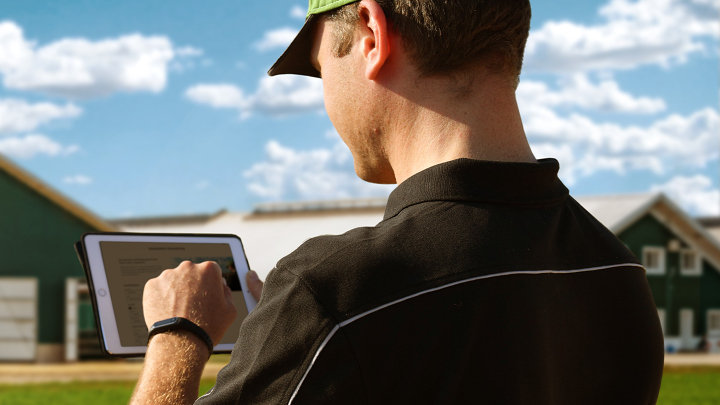 A farmer stands in front of his farm with a tablet in his hand