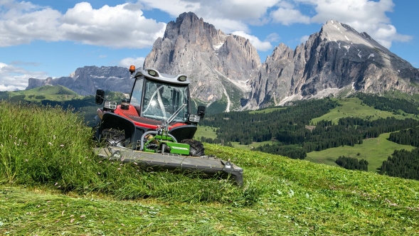 Fendt Slicer Alpine front mower A tractor with the Fendt Slicer Alpine front mower drives on steep terrain and mows a mountain meadow. In the background is a blue sky with white clouds and large mountains.