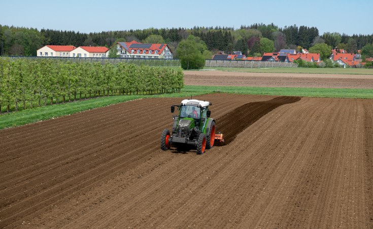 Fendt e100 Vario with ridge tiller A Fendt e107 Vario works with a ridge tiller on a field next to an orchard. In the background, houses with photovoltaic systems on their roofs can be seen.