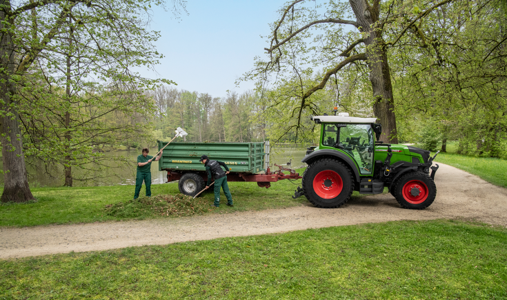 Fendt e100 Vario with a trailer in the park Two landscape conservation workers load a trailer behind a Fendt e107 Vario with leaves in a park with a lake surrounded by trees
